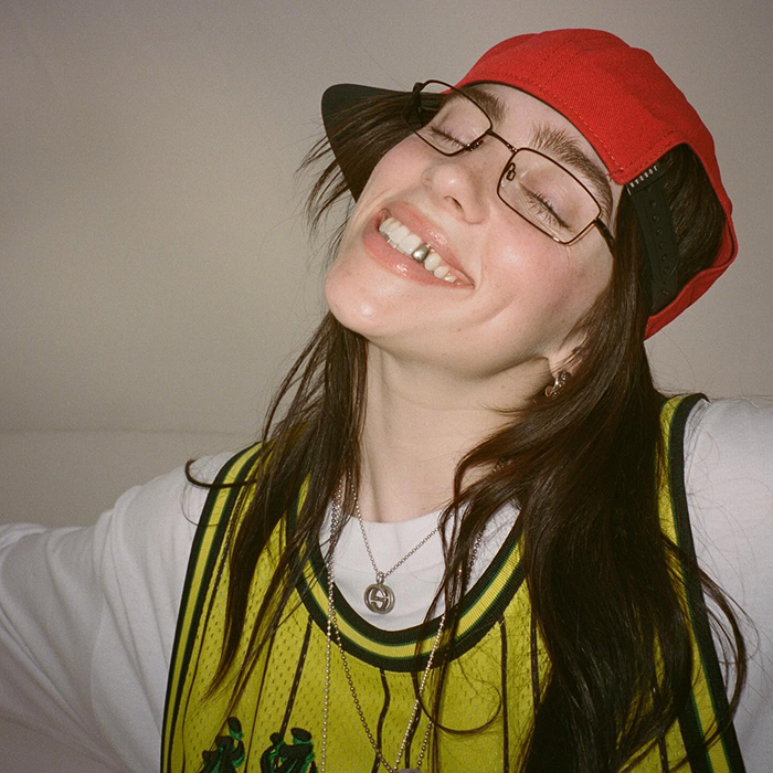 Billie Eilish smiling, wearing a red cap and glasses, reflecting her dating life conversations.