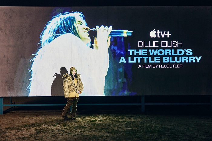 Billie Eilish at film premiere with her image on a billboard, promoting "The World's A Little Blurry.