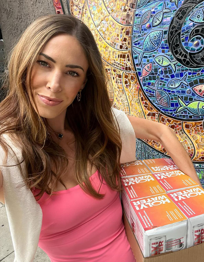 Woman in a pink top holding a box of "Holy Water" against a mosaic wall, relating to American Pie star.