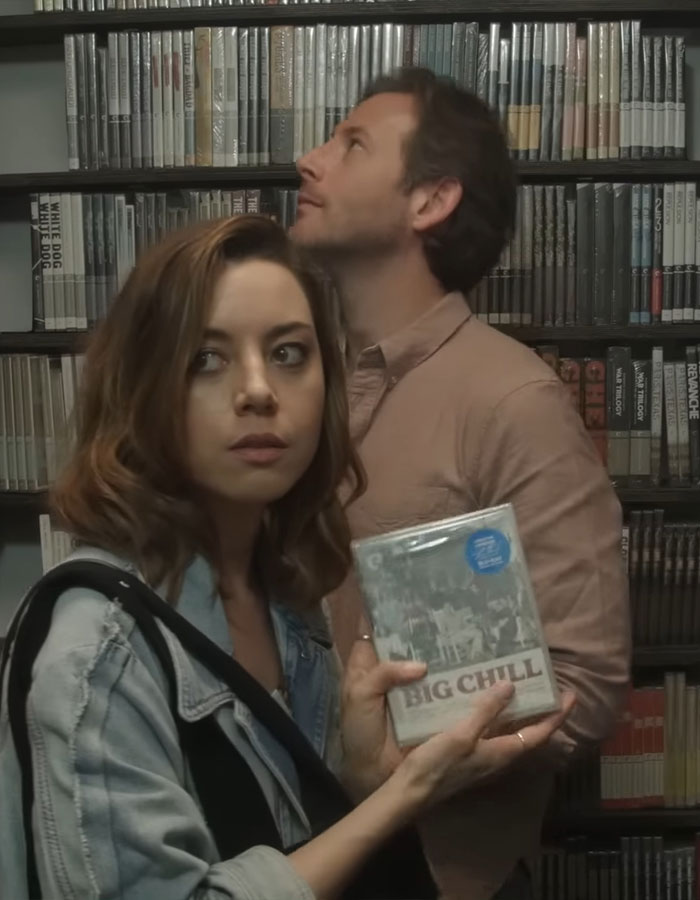 A woman holds a DVD titled "Big Chill" while a man looks upward in a room with shelves.