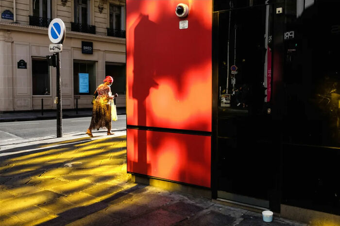A woman with red hair walks past a vivid red wall, capturing the absurd human condition in a colorful urban scene.