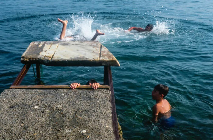 Three boys swimming and diving near a concrete platform, captured by a Belgian photographer embracing the absurd.