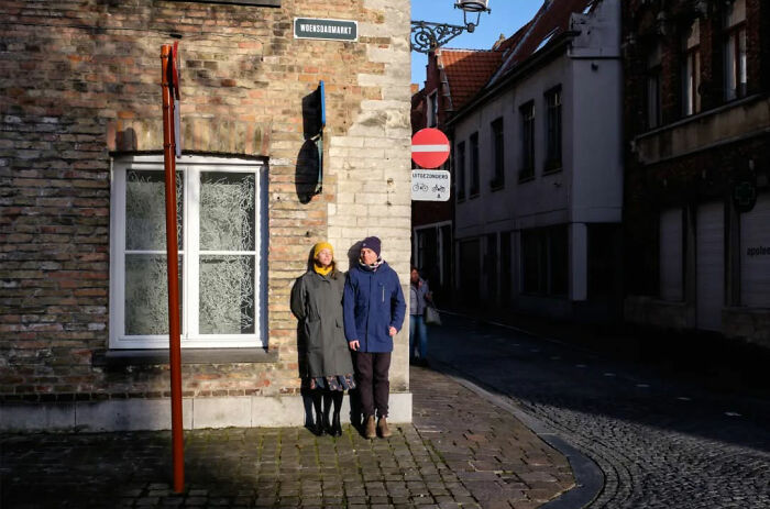 Two people in winter coats standing on a sunlit street corner, embracing the absurd human condition.