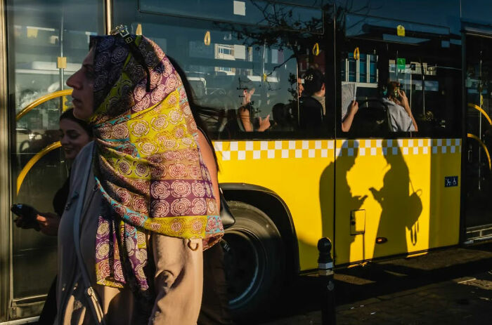 People walking past a yellow bus reflecting shadows, capturing the absurd human condition in a street scene.