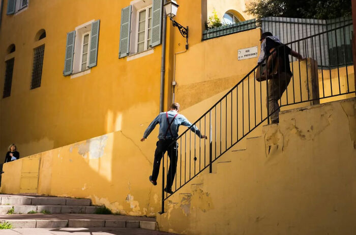 Absurd human condition: man walking up stairs in a peculiar manner against a bright yellow building backdrop.