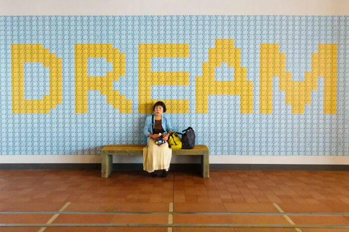 Person seated on a bench in front of a mural with "DREAM," captured by Belgian photographer exploring absurd human condition.