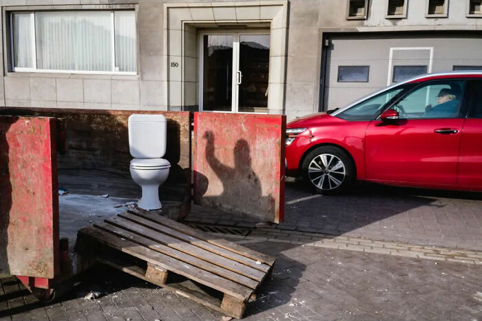 A toilet on a sidewalk next to a red car, casting an unusual shadow.