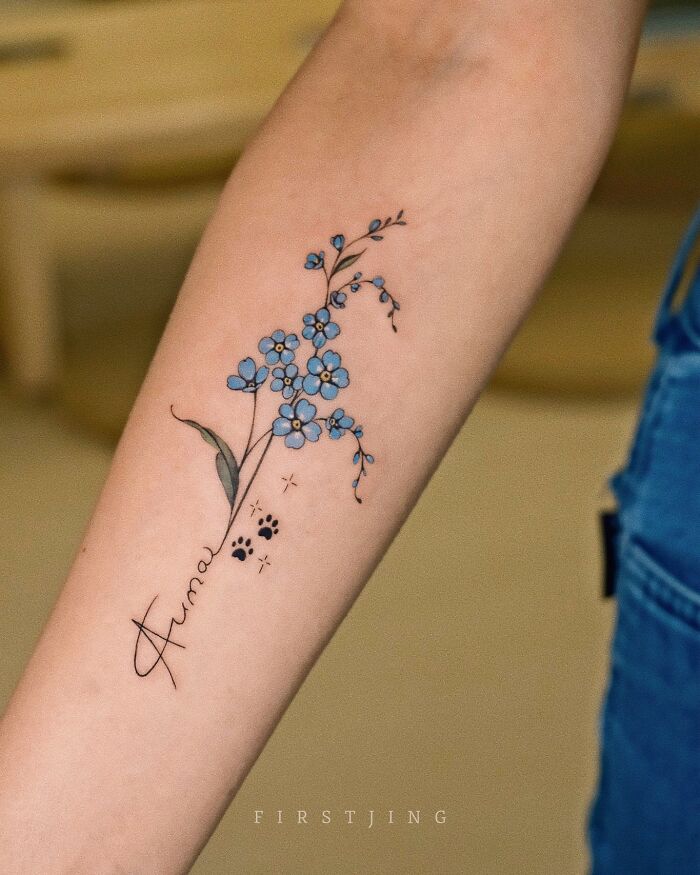 Fine-line tattoo of blue flowers and paw prints on an arm.