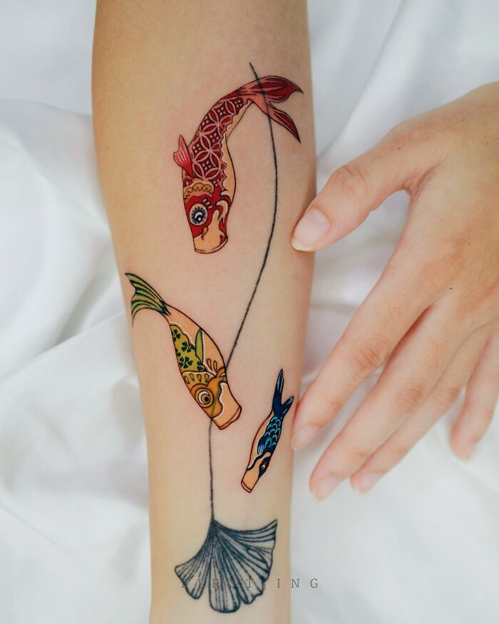 Fine-line tattoos featuring colorful fish and a leaf on an arm.