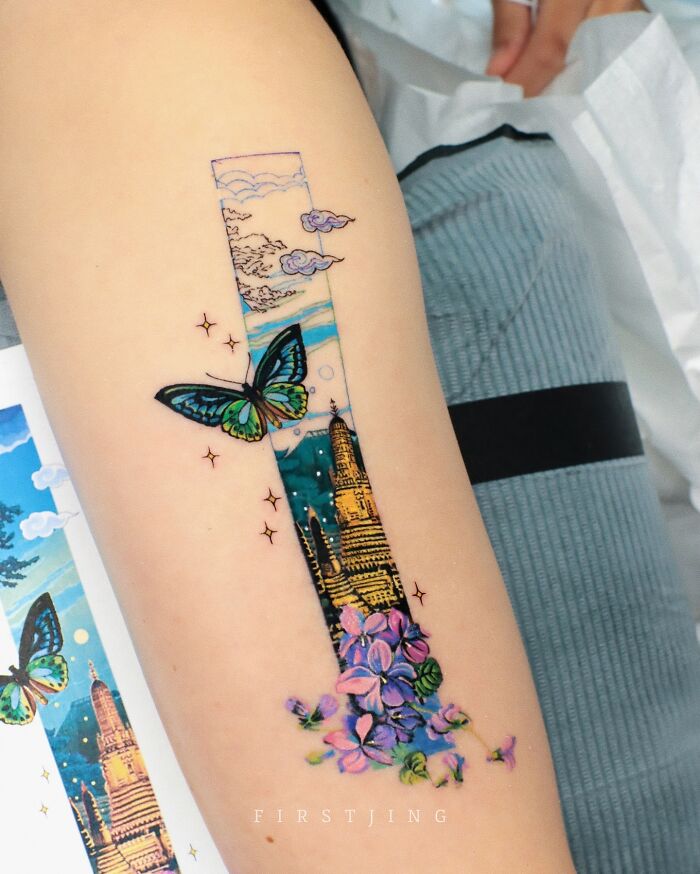 Fine-line tattoo of a butterfly and flowers with dreamy landscape on an arm.