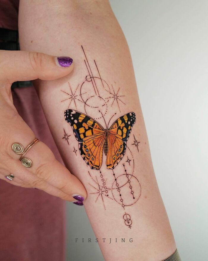 A fine-line tattoo of a butterfly with dreamy settings and geometric patterns on an arm, showcasing artistic detail.
