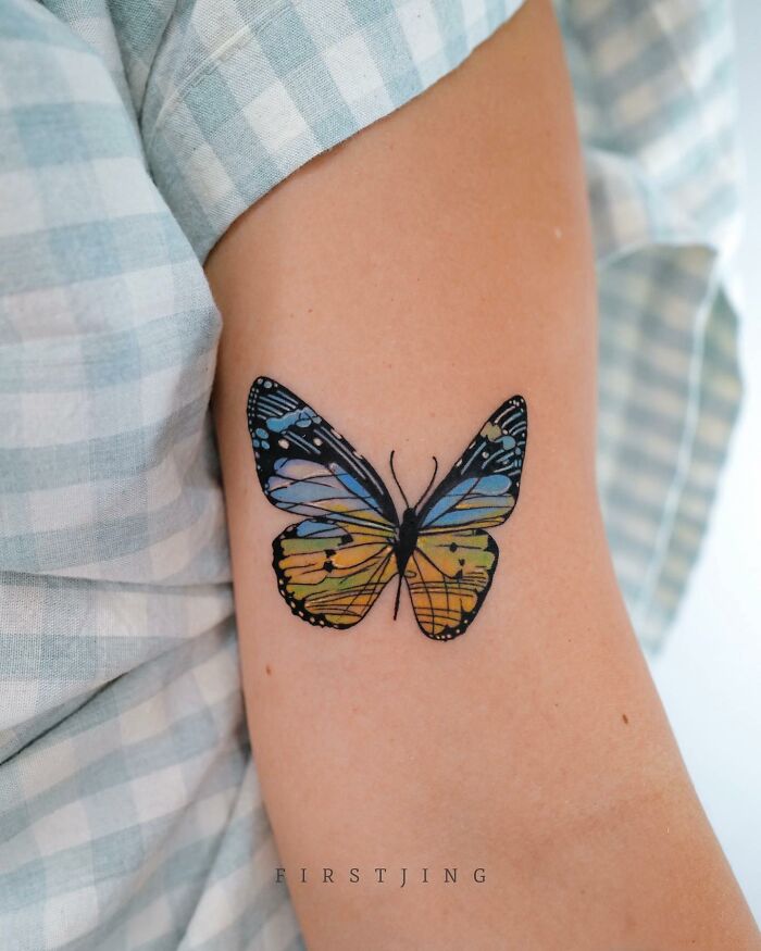 Fine-line tattoo of a colorful butterfly on a person's arm, showcasing dreamy settings and artistic detail.