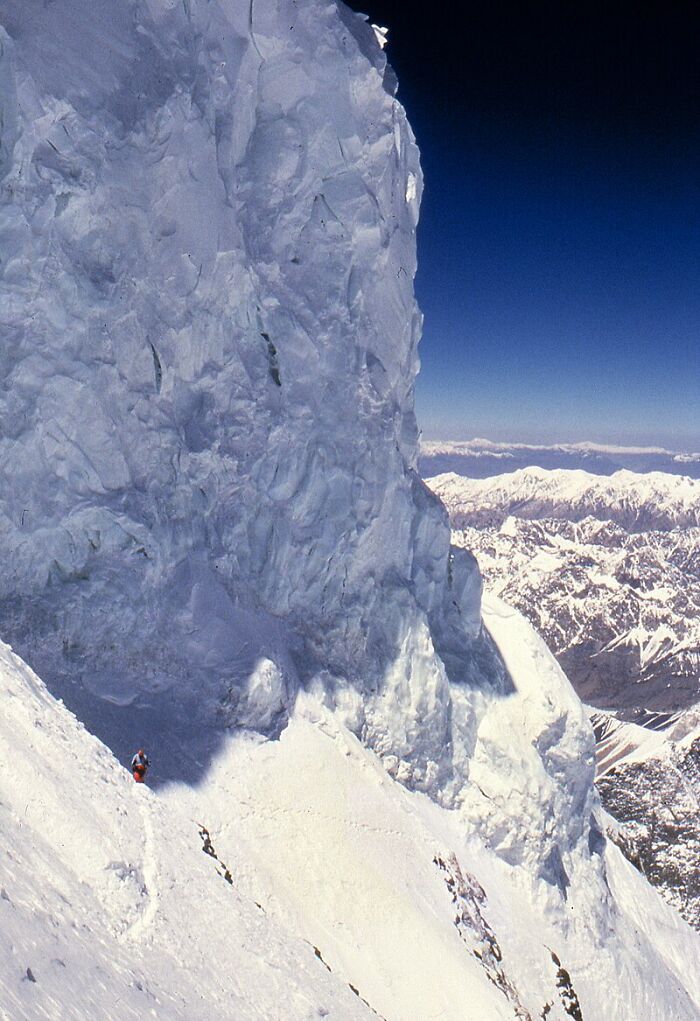 Climber scaling a massive, icy mountain cliff under a deep blue sky, evoking creepy and mysterious historical vibes.