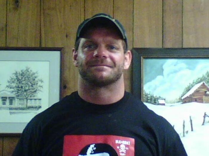 Man in a black shirt and cap standing in front of wooden wall with paintings, related to a creepy photo history.