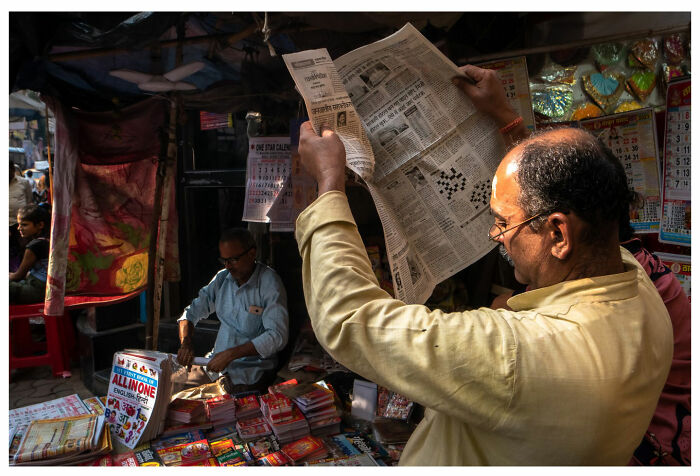 A man reads a newspaper at a busy street market, with a vendor surrounded by colorful goods.