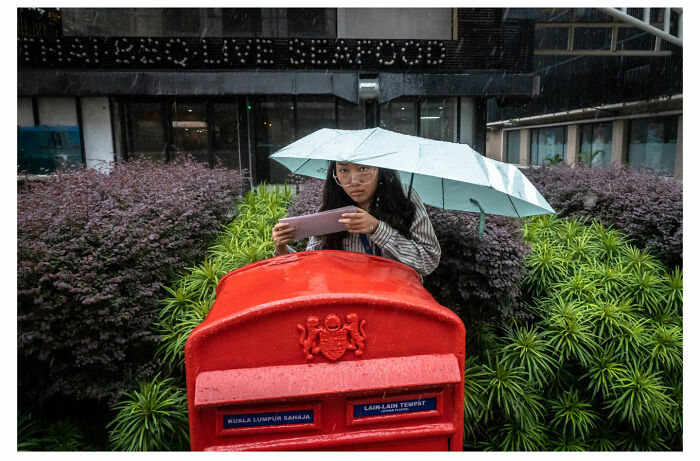 Person with an umbrella and phone near a red mailbox, capturing street moments in rainy weather.