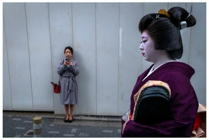 Street photography by Andrés Ramos: a woman in a kimono walks past someone using a phone in an urban setting.
