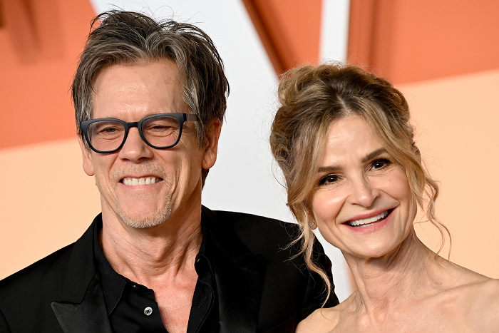 Kyra Sedgwick and Kevin Bacon smiling at a formal event, with the man wearing glasses and a dark suit.