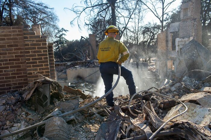 Firefighter in LA County uniform at wildfire site amidst debris and smoke.