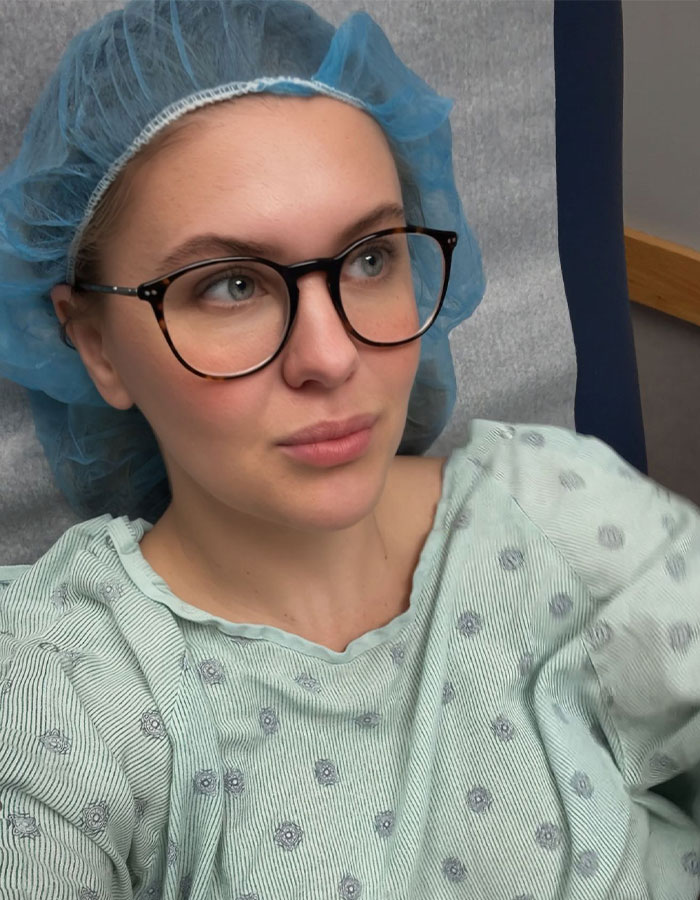Person in a hospital gown and blue cap, wearing glasses, resting on a hospital bed.