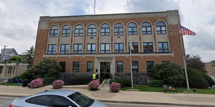 Street view of a police office building with flags, shrubs, and a parked car in front.
