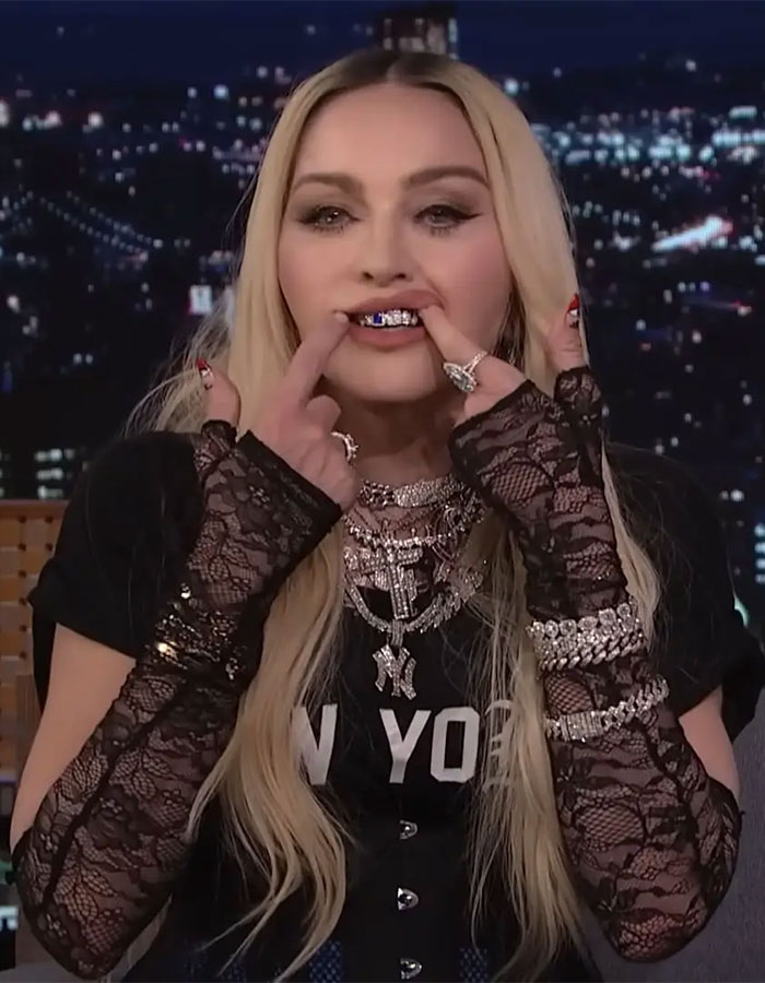 Pop star showcasing diamond grills, wearing lace gloves and layered necklaces, with cityscape in the background.