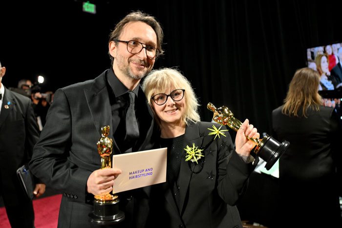 Winners at the Oscars 2025 holding awards and an envelope for Makeup and Hairstyling, smiling on stage.