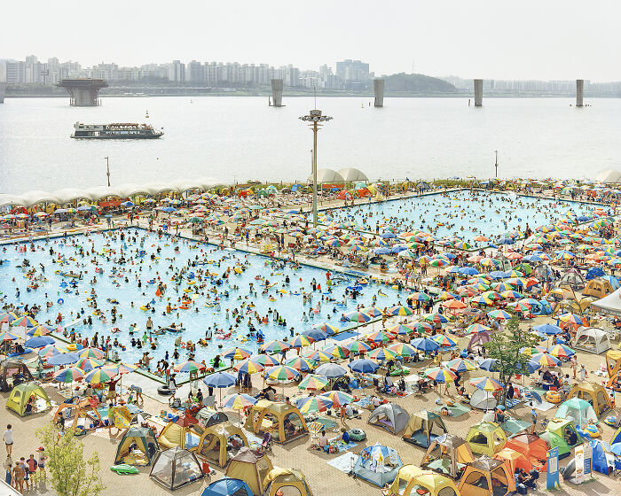 A busy poolside scene with colorful umbrellas and people swimming, set against a city skyline. Stunning photo finalist.