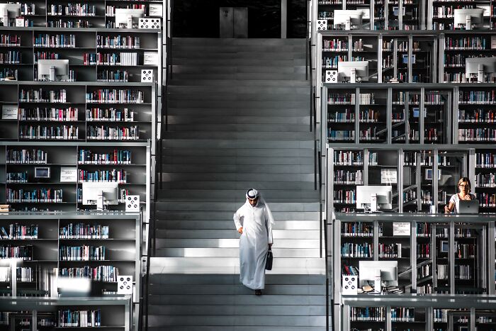 Man in traditional attire walking down library stairs, surrounded by shelves, from stunning 2024 IPA photo collection.