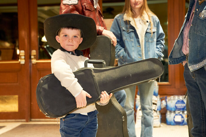 Young boy in a cowboy hat holding a guitar case, surrounded by casually dressed adults. Stunning photography from 2024 IPA.