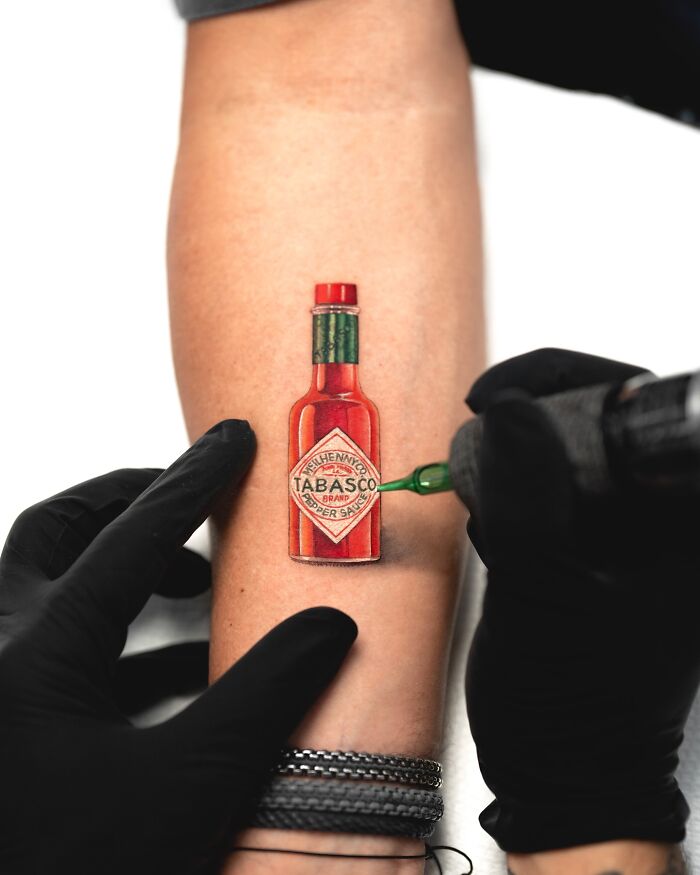 Tattoo of a lifelike Tabasco bottle being inked on an arm, created by Tatu Panda.