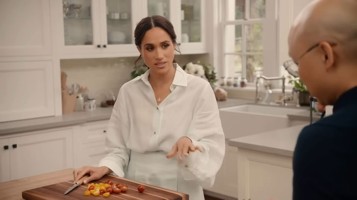 Meghan Markle in a kitchen, discussing with a crew member, tomato slices on a cutting board, wearing a white blouse.