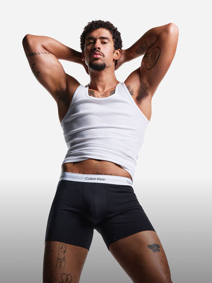 Man in Calvin Klein ad wearing a white tank top and black boxer briefs, showcasing tattoos and posing confidently.