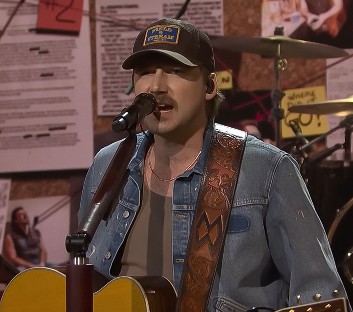 Musician in denim jacket performing on stage with a guitar, drums in the background, focused on singing.