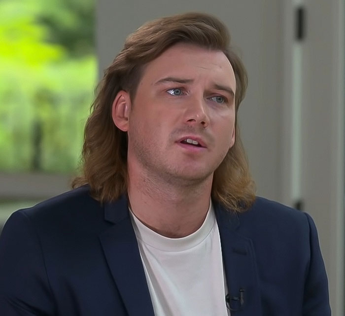 A person in a blue suit jacket, looking thoughtful during an interview, with the spotlight on his reaction on SNL.