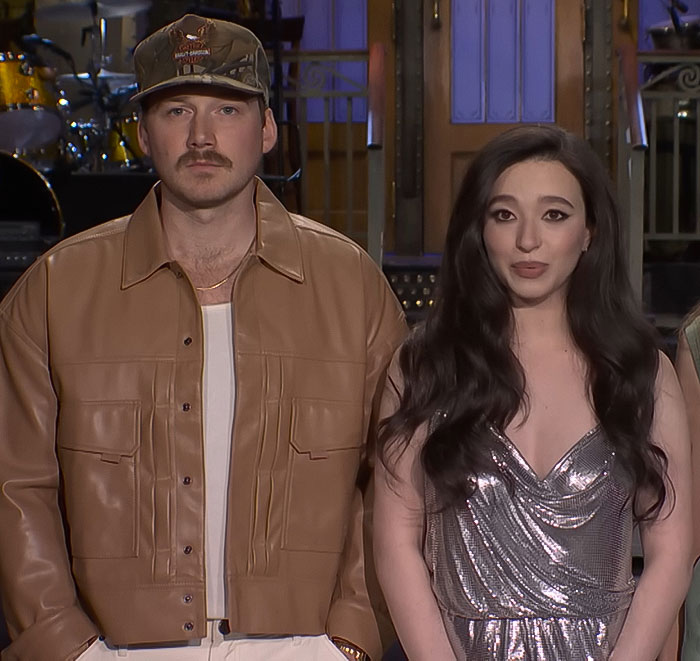Man in a brown jacket and cap next to a woman in a silver dress on stage.