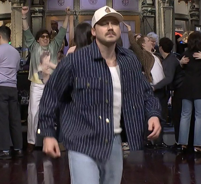 Morgan Wallen on SNL stage, wearing a striped jacket and cap, with a visible crowd in the background.