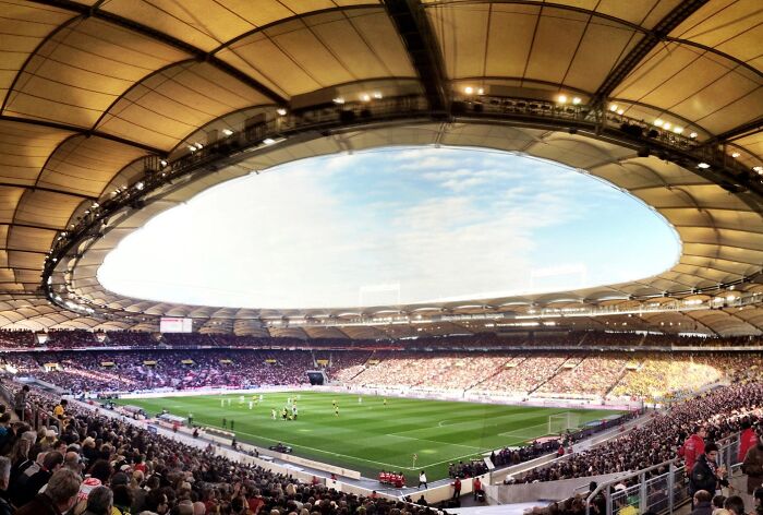 Panoramic view of a packed soccer cathedral stadium with vibrant fans and players on the green pitch under a bright sky.