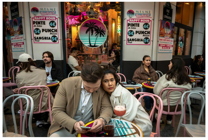 Couple sitting closely at outdoor café, surrounded by people, capturing raw, unfiltered street moments with vibrant colors.