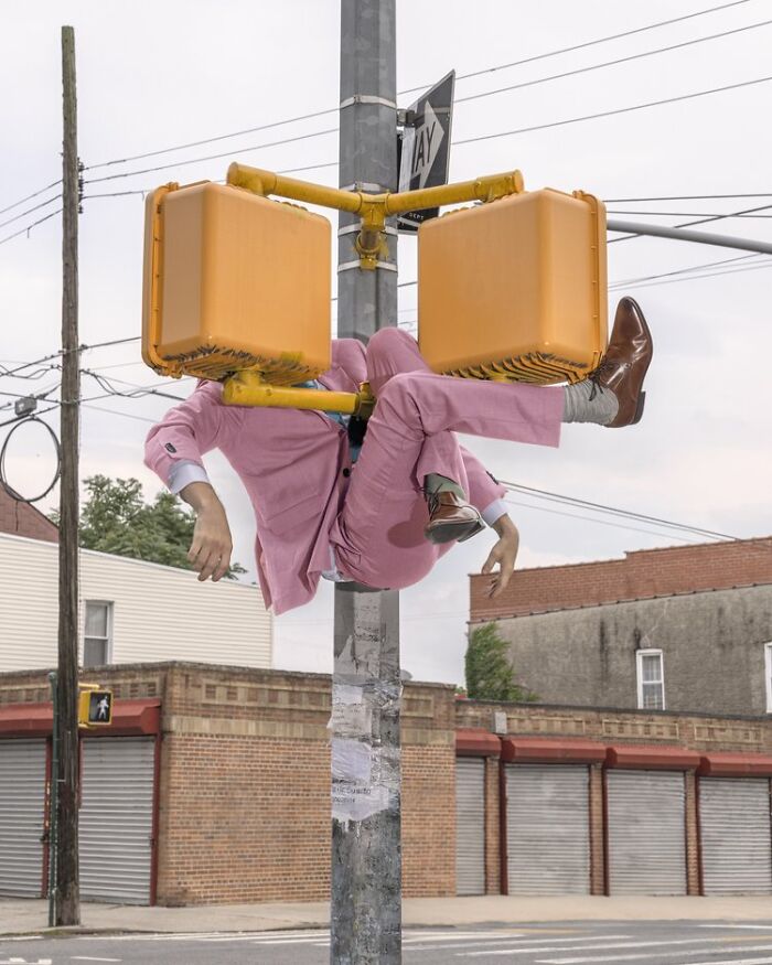 Solitary figure in a pink suit, hanging unusually from a streetlight, showcasing surreal photography by Ben Zank.
