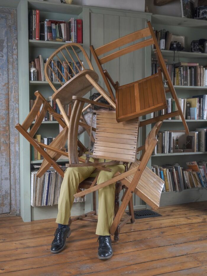 Solitary figure buried under stacked chairs in a room with wooden floors and bookshelves, illustrating surreal photography.