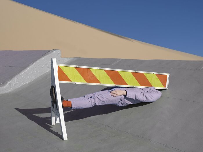 Surreal photograph by Ben Zank of a solitary figure in a unique pose under a road barrier in a minimalistic landscape.