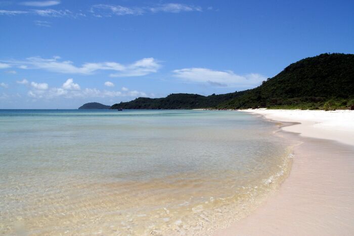 Stunning beach with clear water and white sand, surrounded by lush green hills under a bright blue sky.