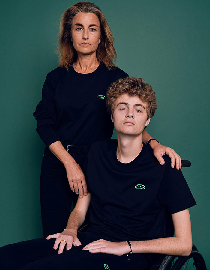 Woman standing beside a young man in a wheelchair against a green background, both wearing black shirts. Woman standing beside a young man in a wheelchair against a green background, both wearing black shirts.