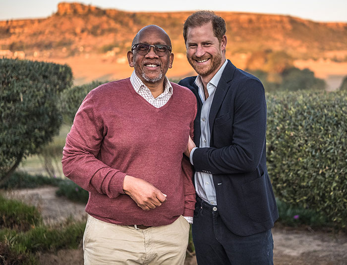 Two men smiling outdoors with greenery and sunset, related to Prince Harry and charity.