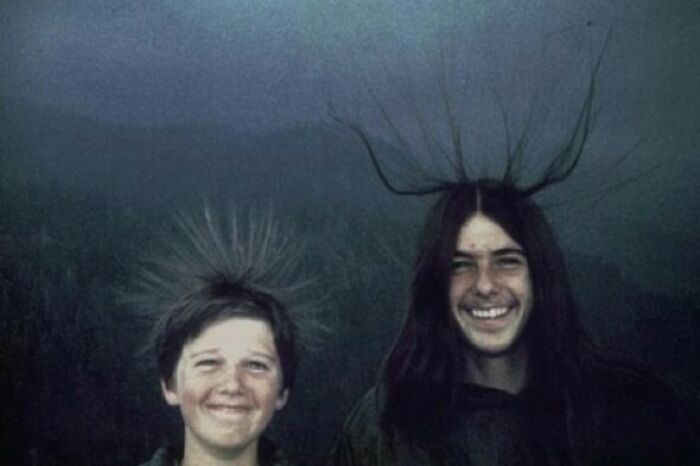 Two people smiling with static hair standing outdoors, under dark clouds, evoking a creepy atmosphere.