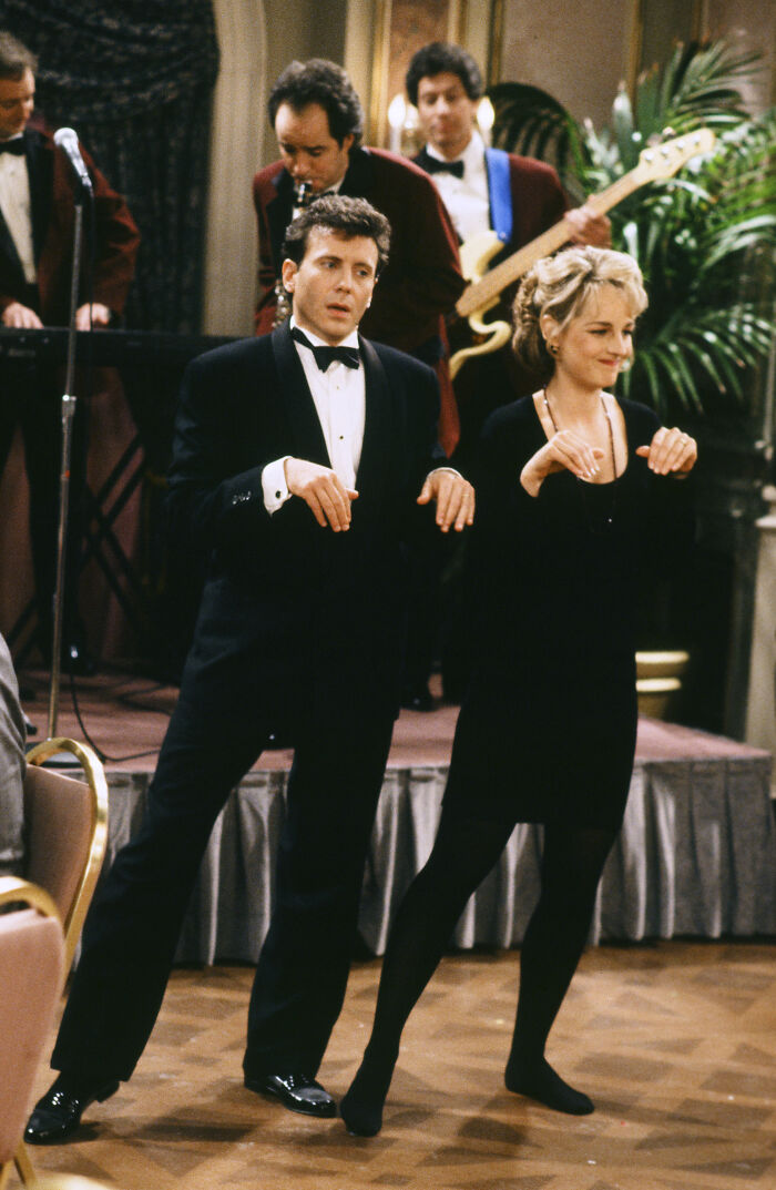 A woman in a black outfit mimics dance moves beside a man in a tuxedo at a lively indoor event. A woman in a black outfit mimics dance moves beside a man in a tuxedo at a lively indoor event.