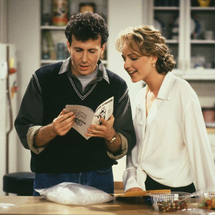 A woman in a white blouse reads alongside a man holding a booklet in a kitchen setting. A woman in a white blouse reads alongside a man holding a booklet in a kitchen setting.