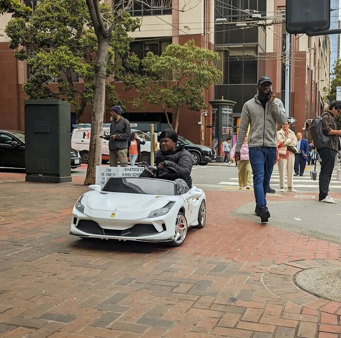 Street scene in San Francisco with people walking past a person riding a small, white toy car on the sidewalk.