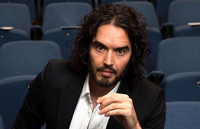 A person with long curly hair and a beard, wearing a black blazer, sits in a theater, showcasing a more attractive presence in person.